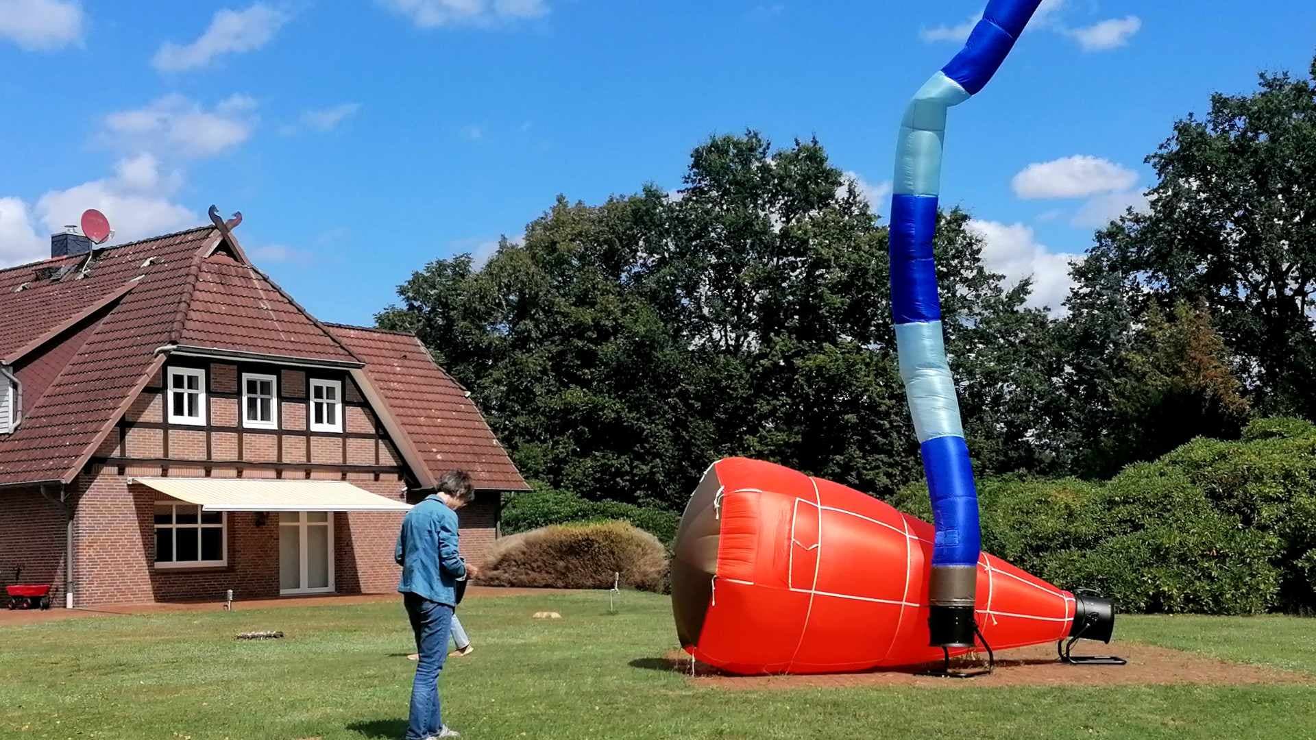 Rote Luftskulptur mit blauem Schlauch im Garten vor einem Fachwerkhaus.
