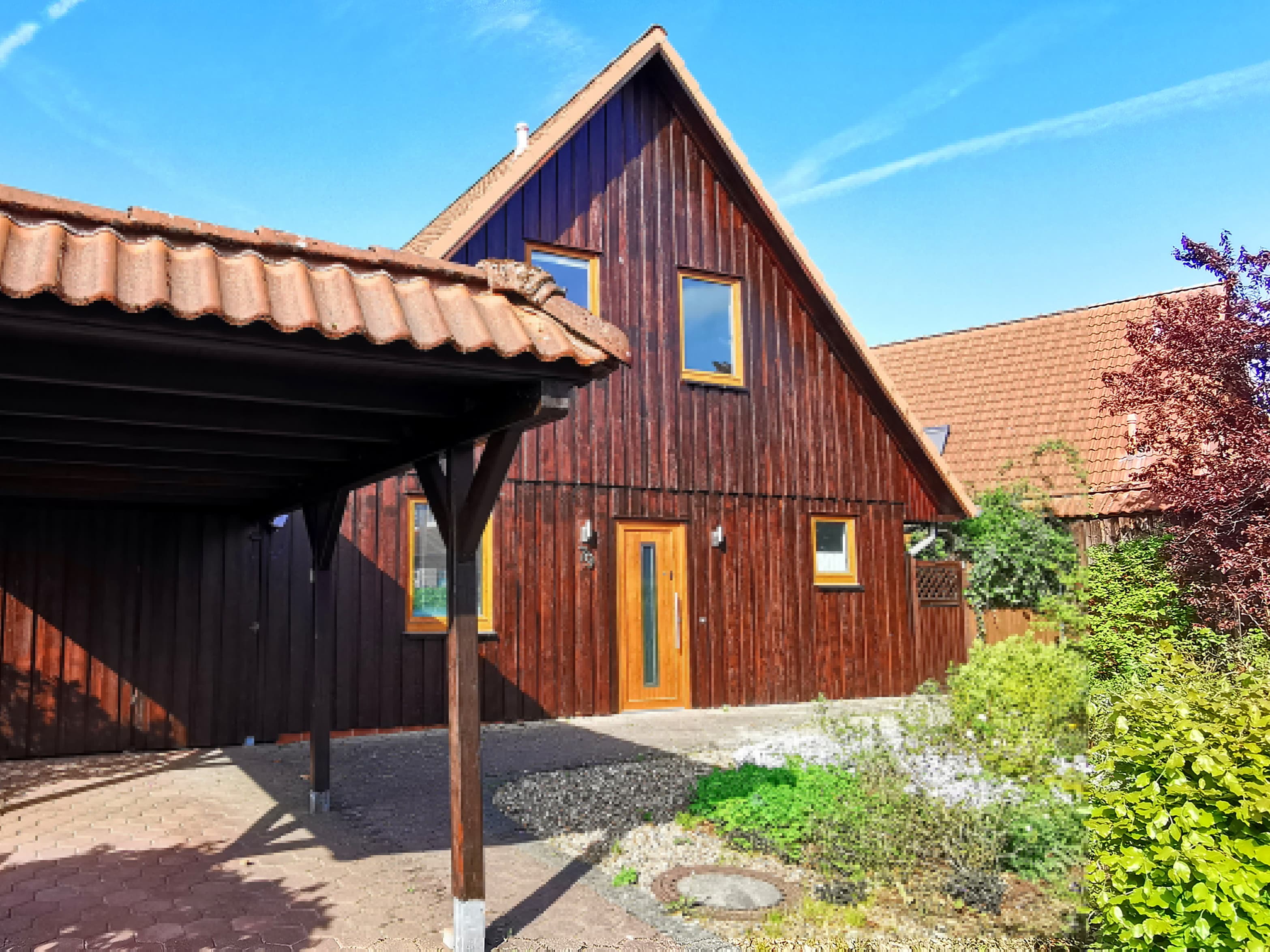 Ein charmantes Holzhaus mit einem steilen, gedeckten Dach. Der Garten ist grün und einladend, und der Himmel ist klar und blau.