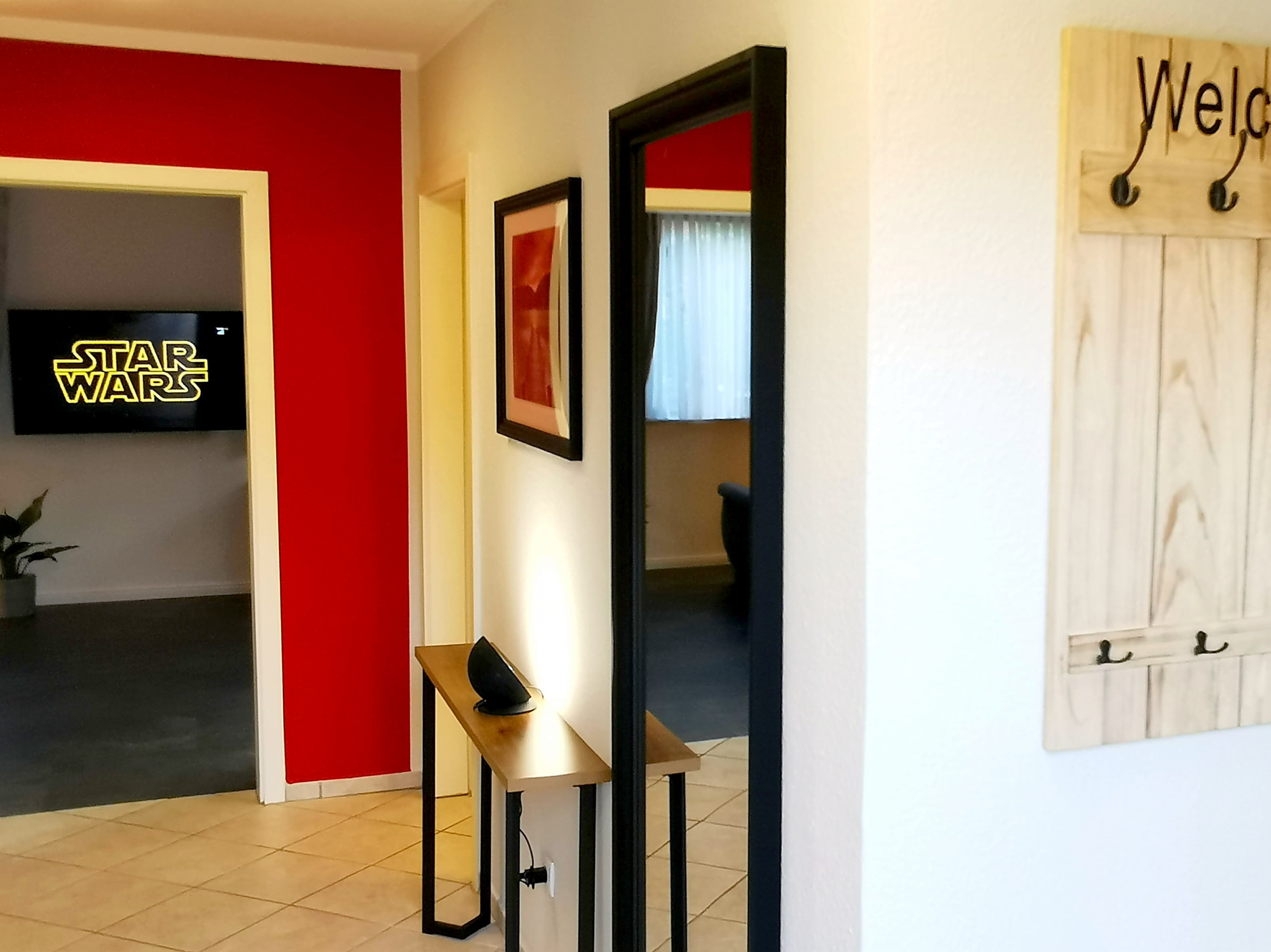 Ein einladender Eingangsbereich mit einem Spiegel und einem Holztablett. An der Wand hängt ein Willkommensschild, und im Hintergrund ist ein Fernseher mit dem Logo von Star Wars zu sehen.