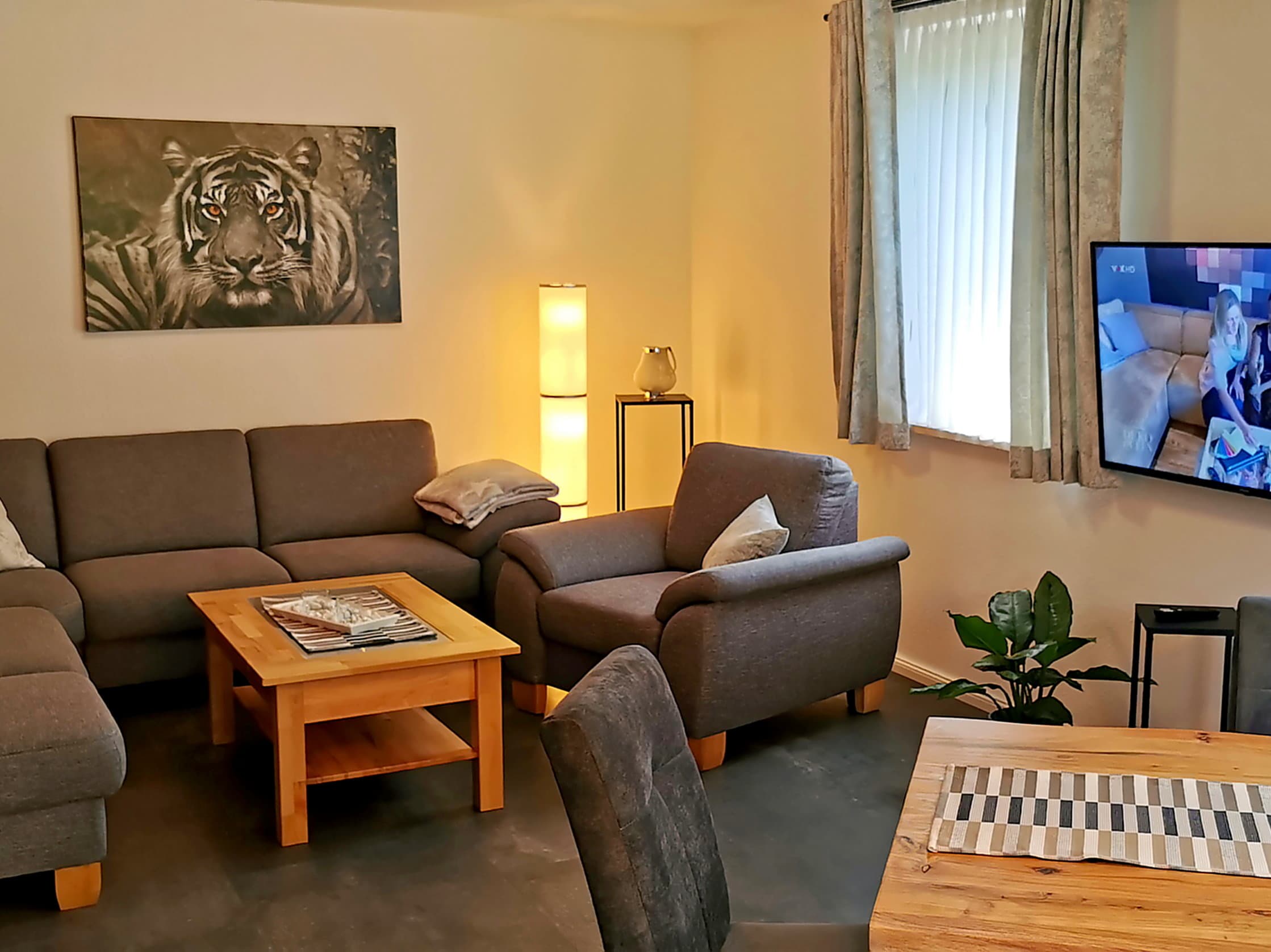 Ein gemütliches Wohnzimmer mit einem grauen Sofa und einem Holztisch. An der Wand hängt ein Bild eines Tigers und es gibt einen Fernseher im Raum.