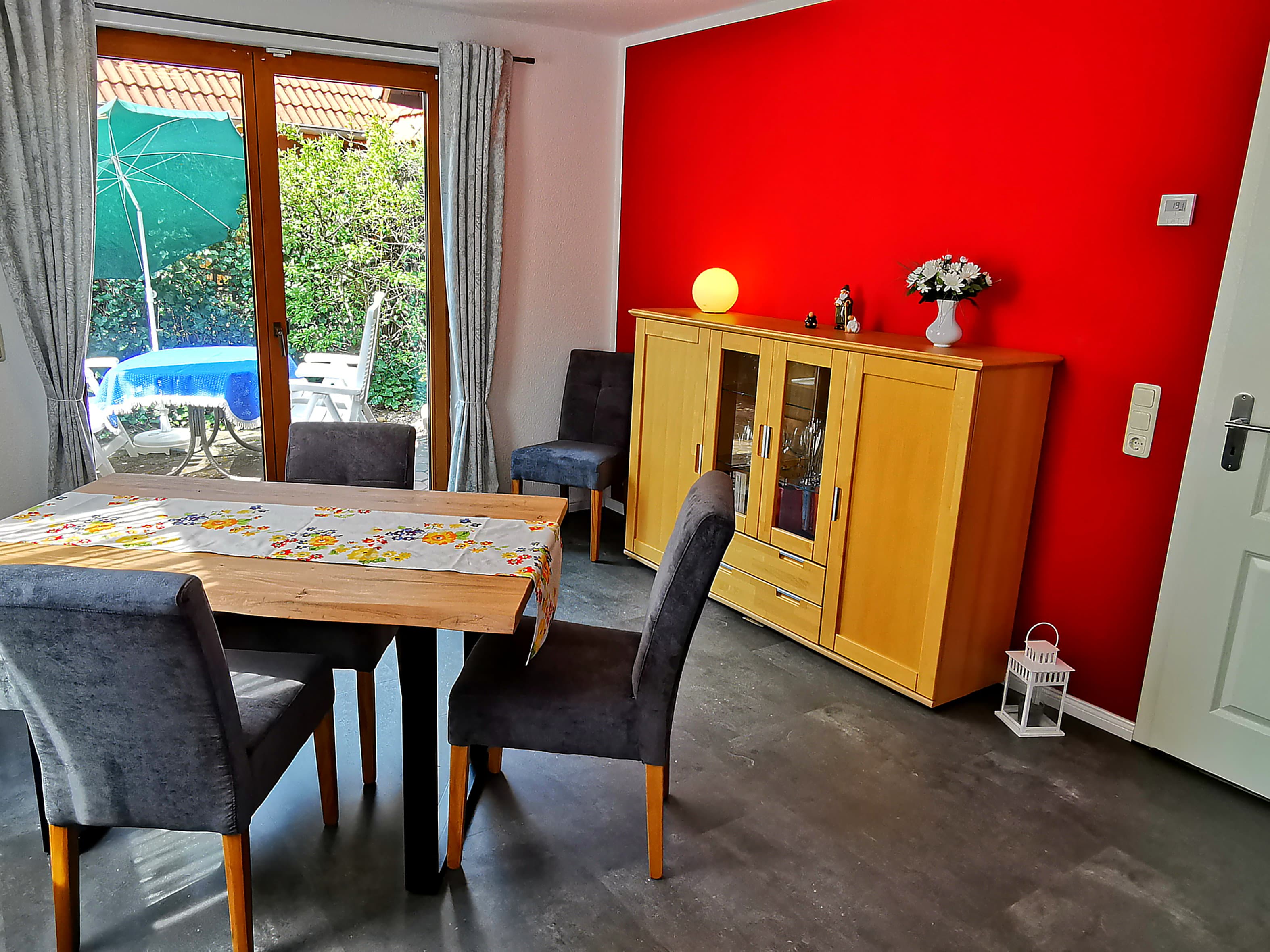 Ein gemütlicher Essbereich mit einem Holztisch und grauen Stühlen. Die Wand ist rot gestrichen, und es gibt Zugang zum Garten.