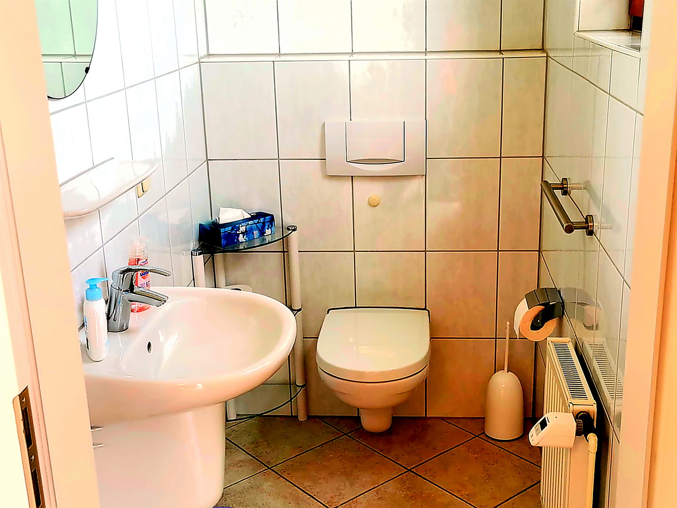 Ein kleines Badezimmer mit weißen Fliesen und einer hellen Atmosphäre. Es gibt ein Waschbecken, eine Toilette und einige Hygieneartikel.