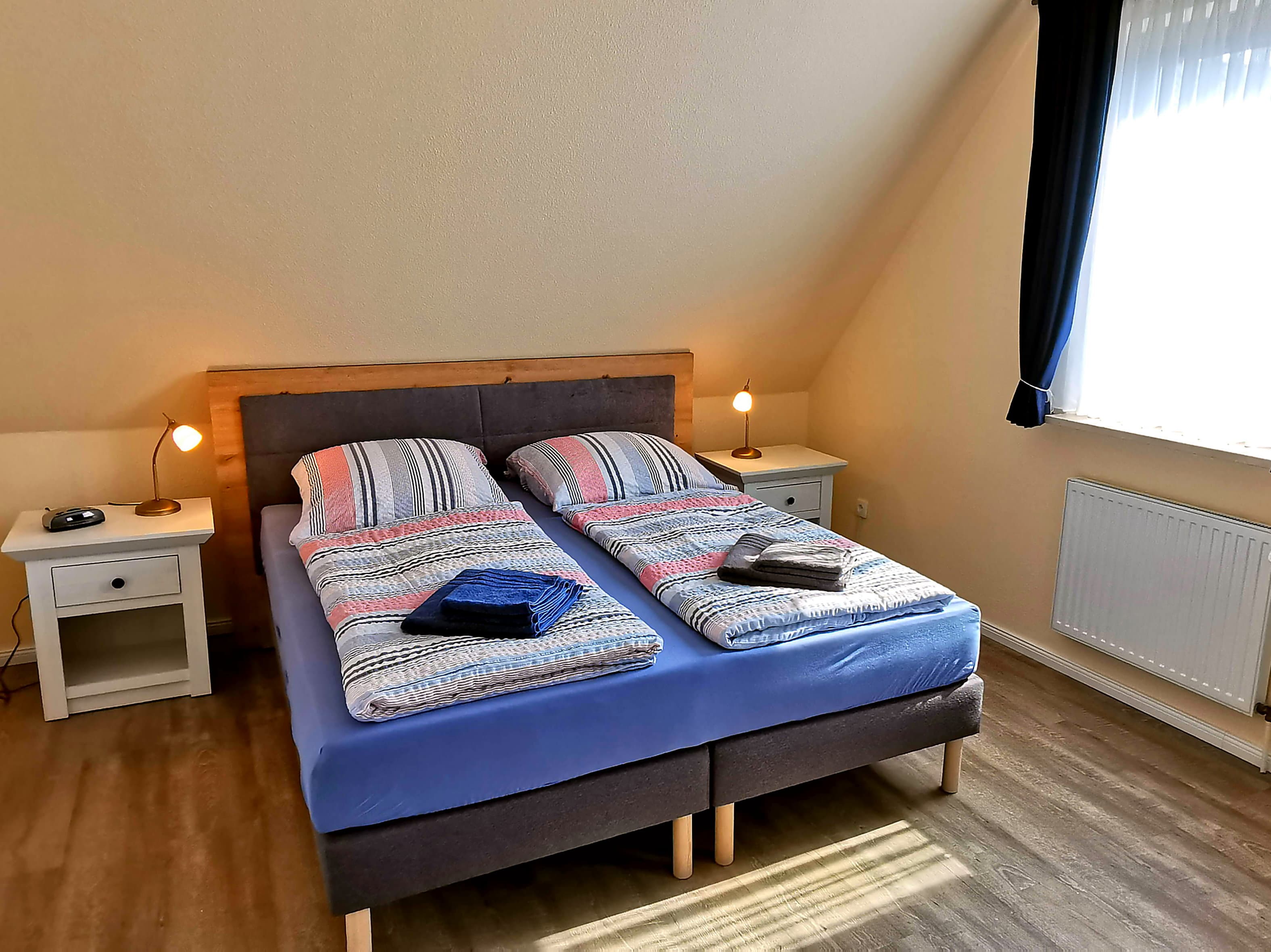Ein helles Schlafzimmer mit einem Doppelbett und gemütlicher Bettwäsche. Auf Nachtkästchen stehen Lampen, und das Zimmer hat große Fenster mit Vorhängen.