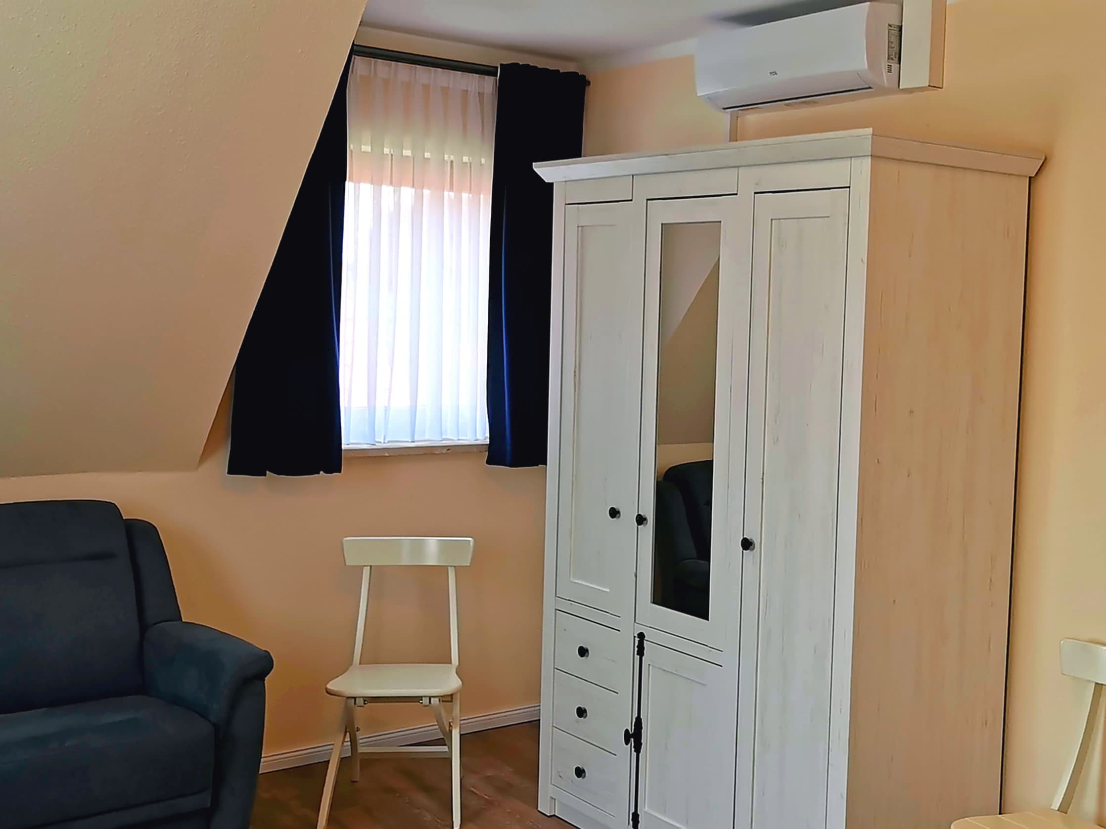Ein helles Zimmer mit einem blauen Sessel und einer weißen Kommode. Die Fenster sind mit weißen Vorhängen und dunklen Gardinen dekoriert.