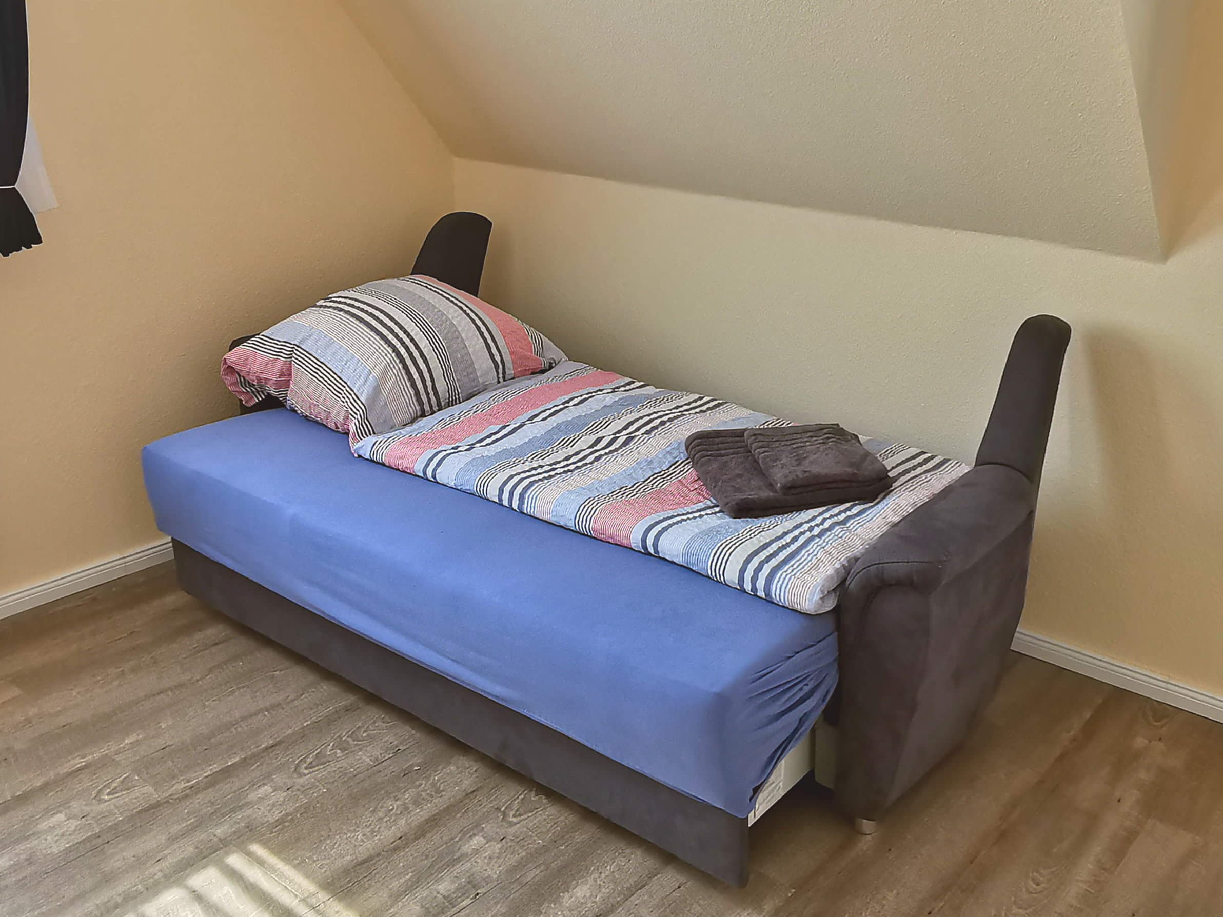 Ein einfaches Bett mit bunten Bettzeug steht in einem Zimmer. Der Boden ist aus Holz, und die Wände sind hell gestrichen.