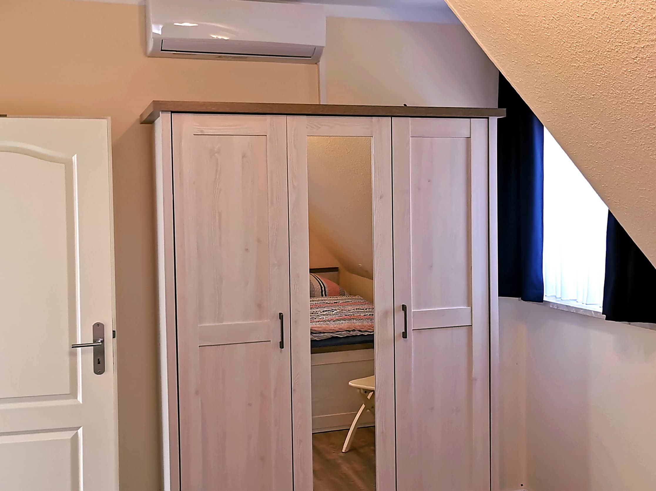 Ein helles Zimmer mit einem modernen Kleiderschrank und einem Bett im Hintergrund. Das Zimmer hat ein Fenster mit Vorhang und eine Klimaanlage an der Wand.