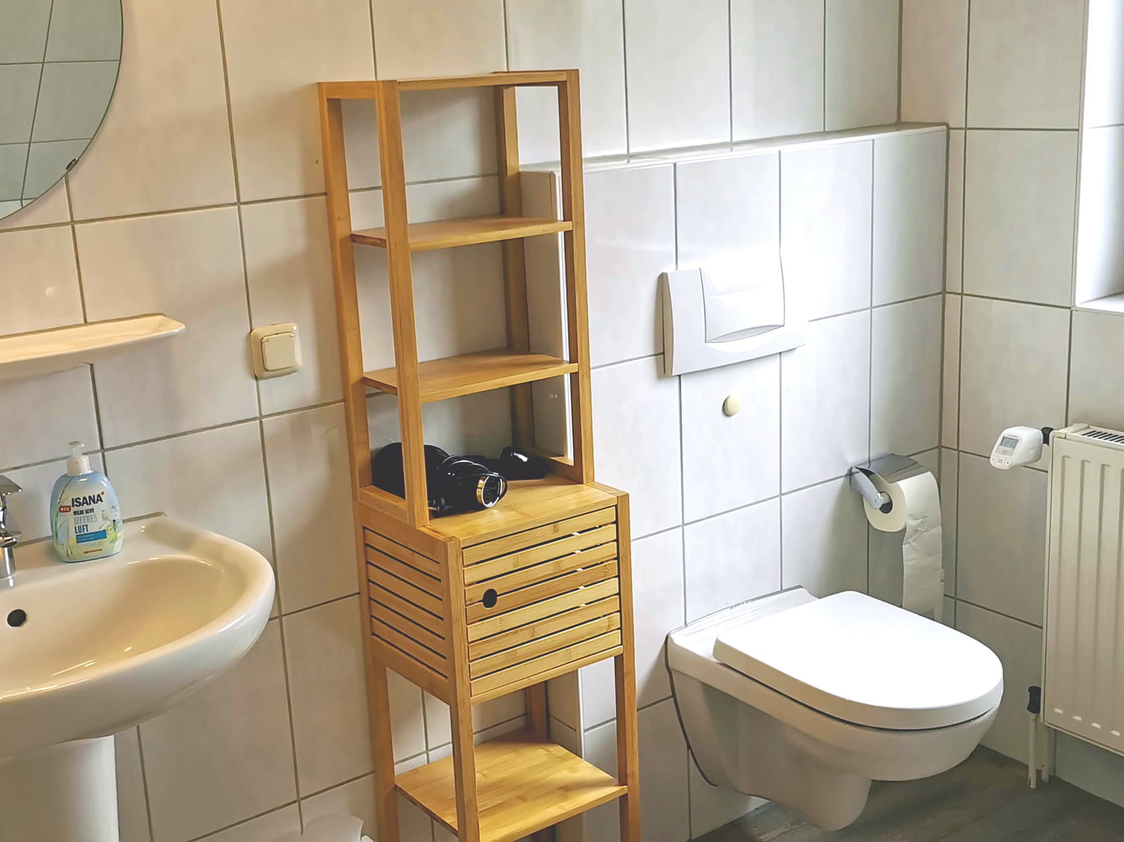 Ein modernes Badezimmer mit einer hellen Fliesenwand. Ein Holzregal steht neben dem Waschbecken und der Toilette.