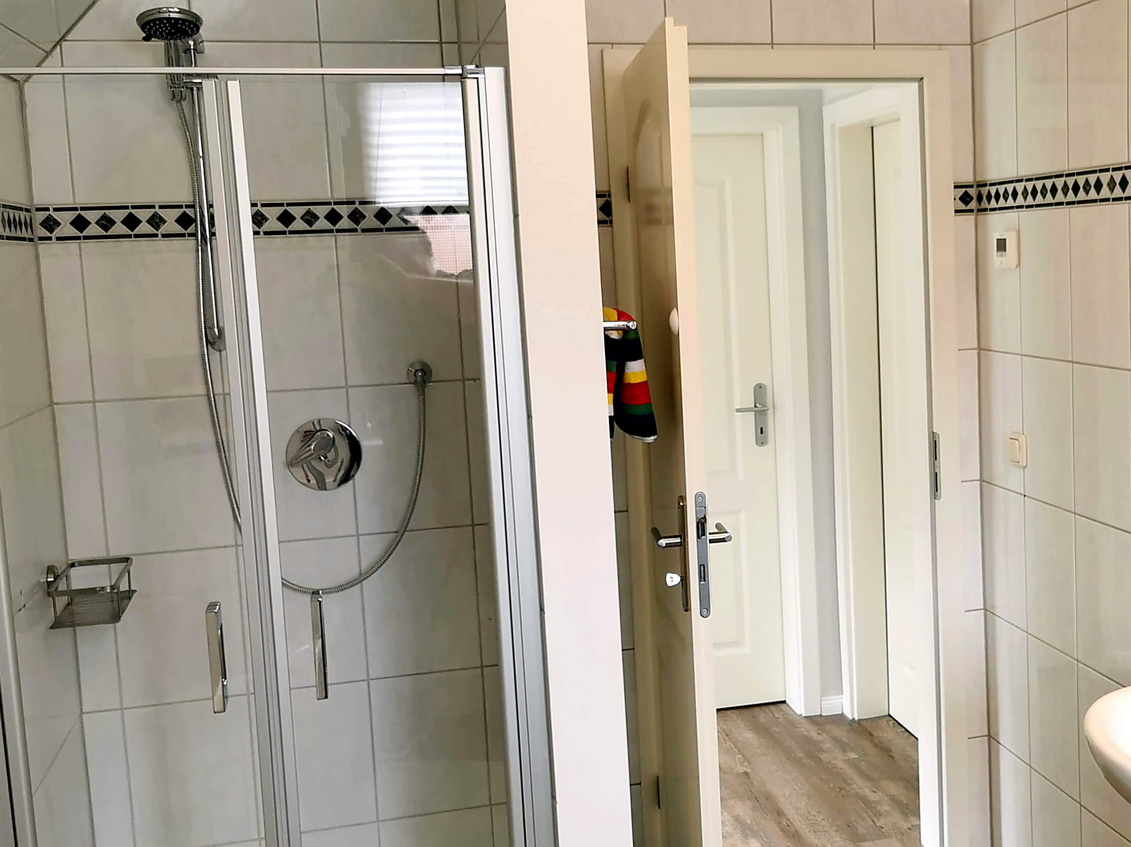 Ein modernes Badezimmer mit einer Dusche und weißer Fliese. Eine geöffnete Tür führt in einen weiteren Raum.