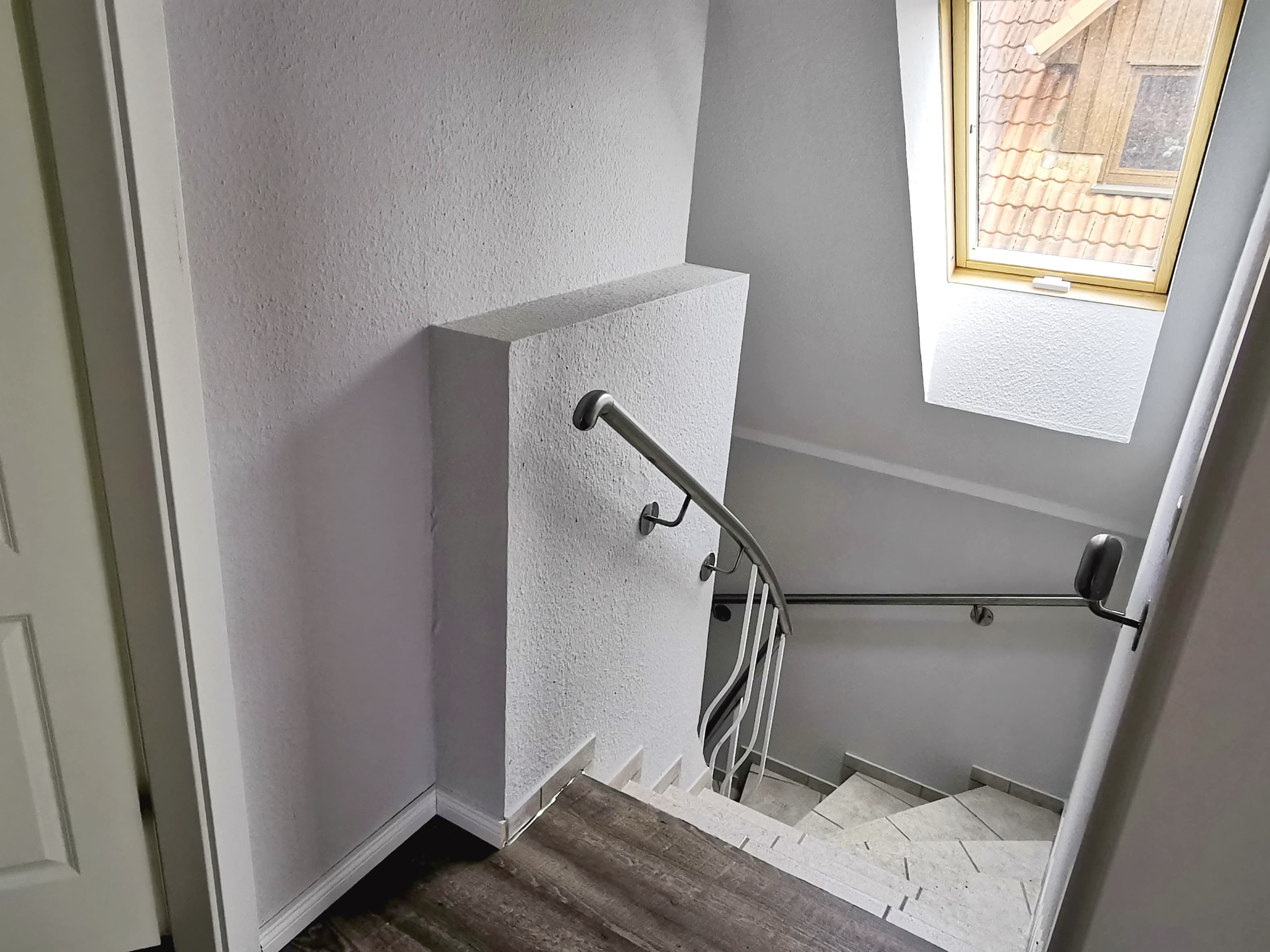Eine Treppe mit grauen Wänden und einem Fenster, das Tageslicht hereinlässt. Der Boden ist mit modernen Fliesen und Holzbelag versehen.