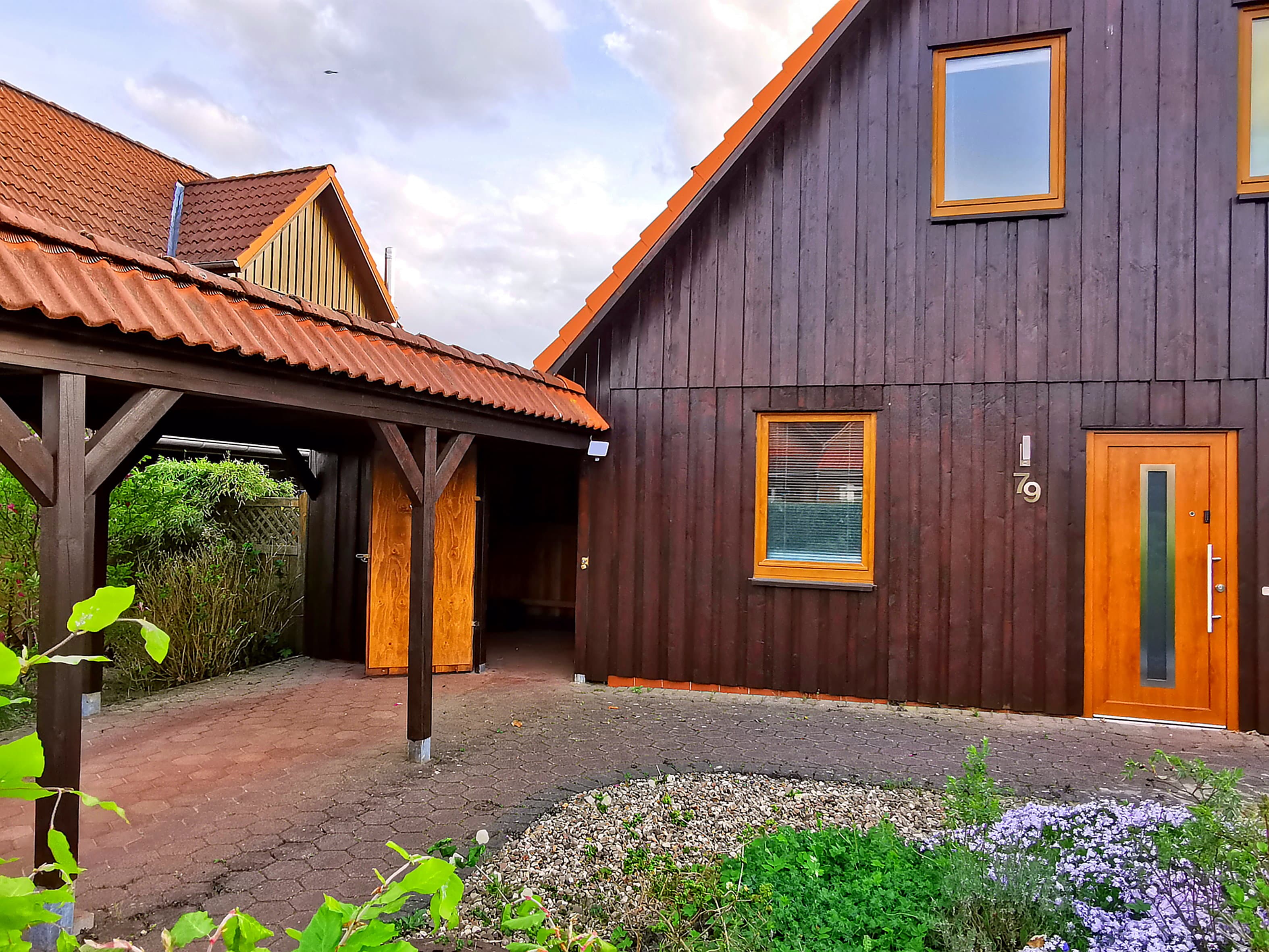 Ein modernes Holzhaus mit einem roten Dach und großen Fenstern. Der Eingang ist von einem gepflasterten Weg und grünen Pflanzen umgeben.