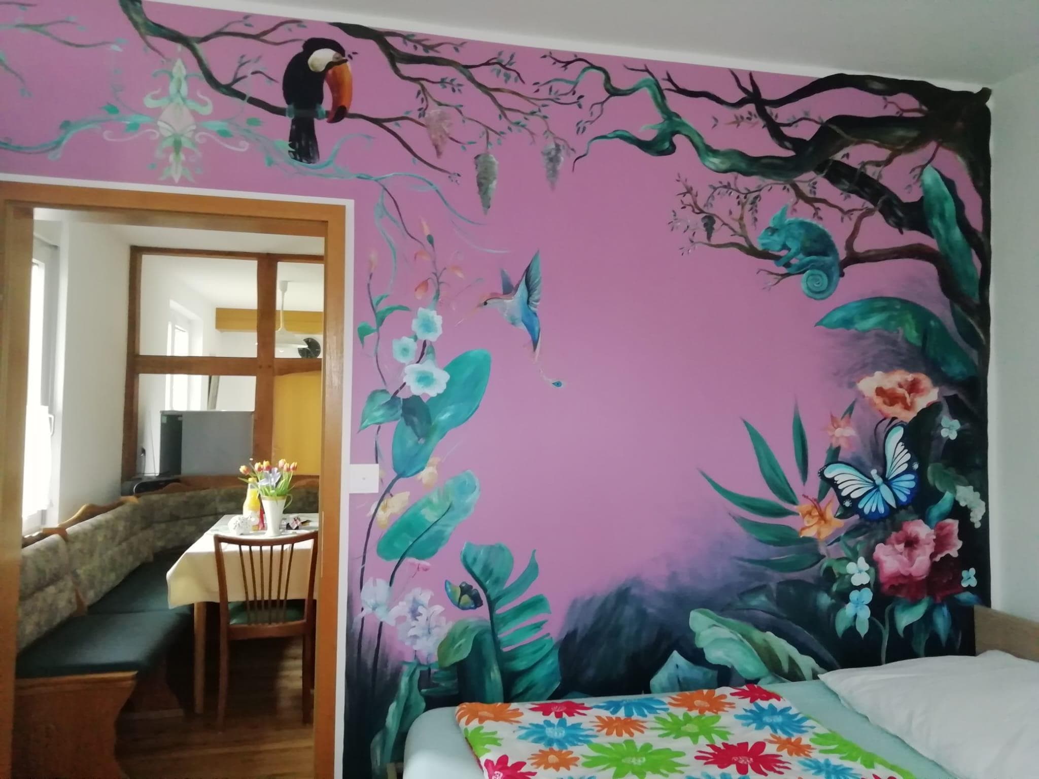 Bett vor rosa Wandmalerei mit tropischen Pflanzen, Tukan, Kolibri und Schmetterling.