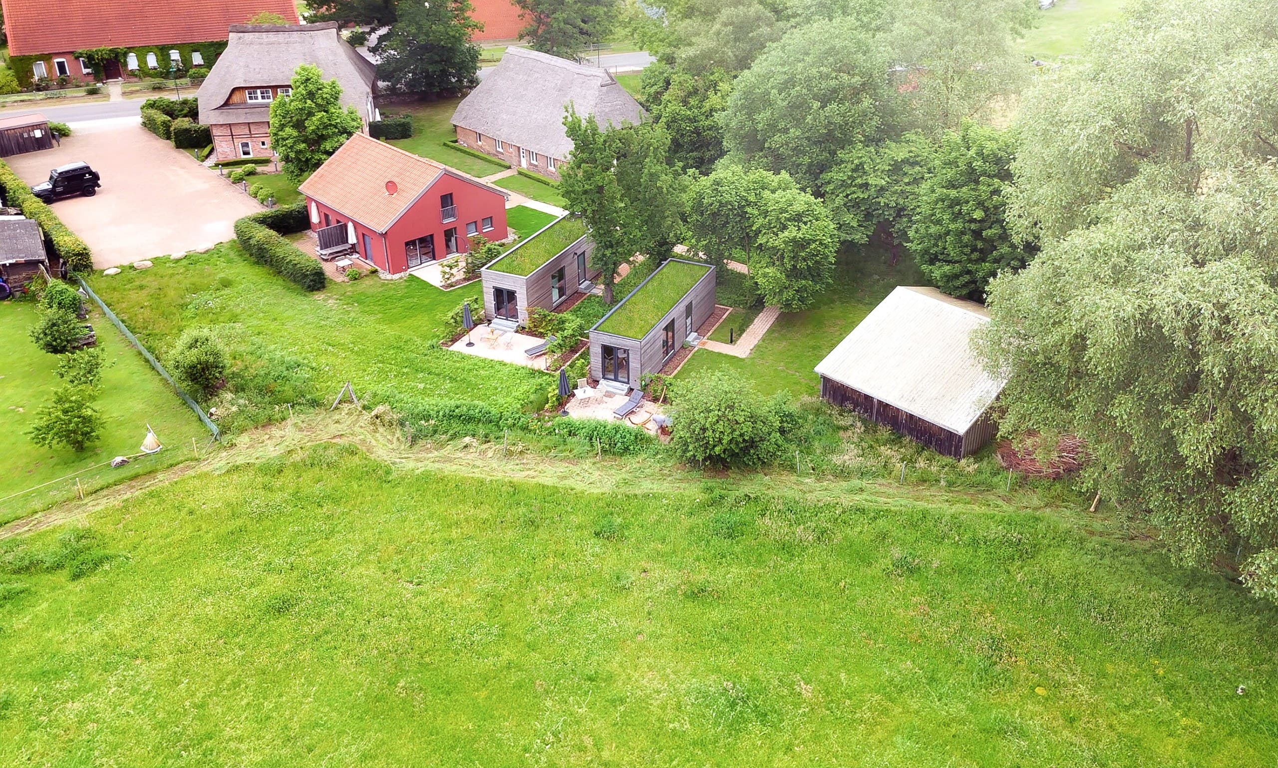 Luftaufnahme des HofGut Bockelkathen mit reetgedeckten Hofgebäuden, rotem Apartmenthaus, zwei Tiny Houses mit Gründach und Wiese
